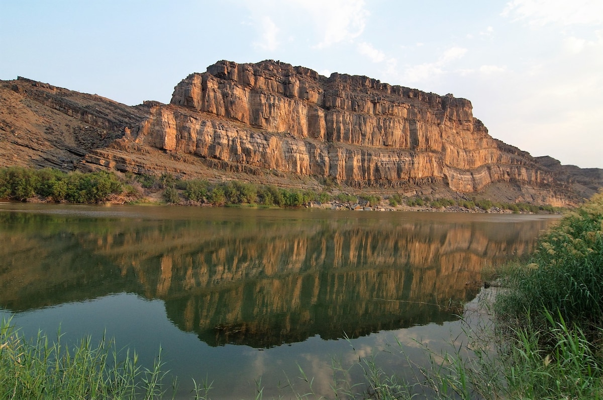 A scenic landscape showcases the rugged cliffs reflecting on the calm waters of the Orange River. Lush greenery lines the riverbank, contrasting with the earthy tones of the rocky formations. The sky is softly illuminated, hinting at the beauty of the surrounding natural environment.