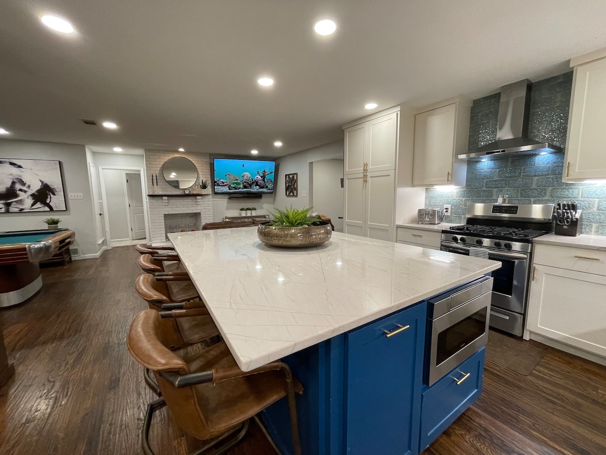 The kitchen showcases a large island with a blue base and grey countertop, accompanied by four metal bar stools. A stainless steel stove is situated alongside white cabinetry, while contrasting blue tiles are featured on the backsplash. A television is mounted on the wall, enhancing the kitchen space.