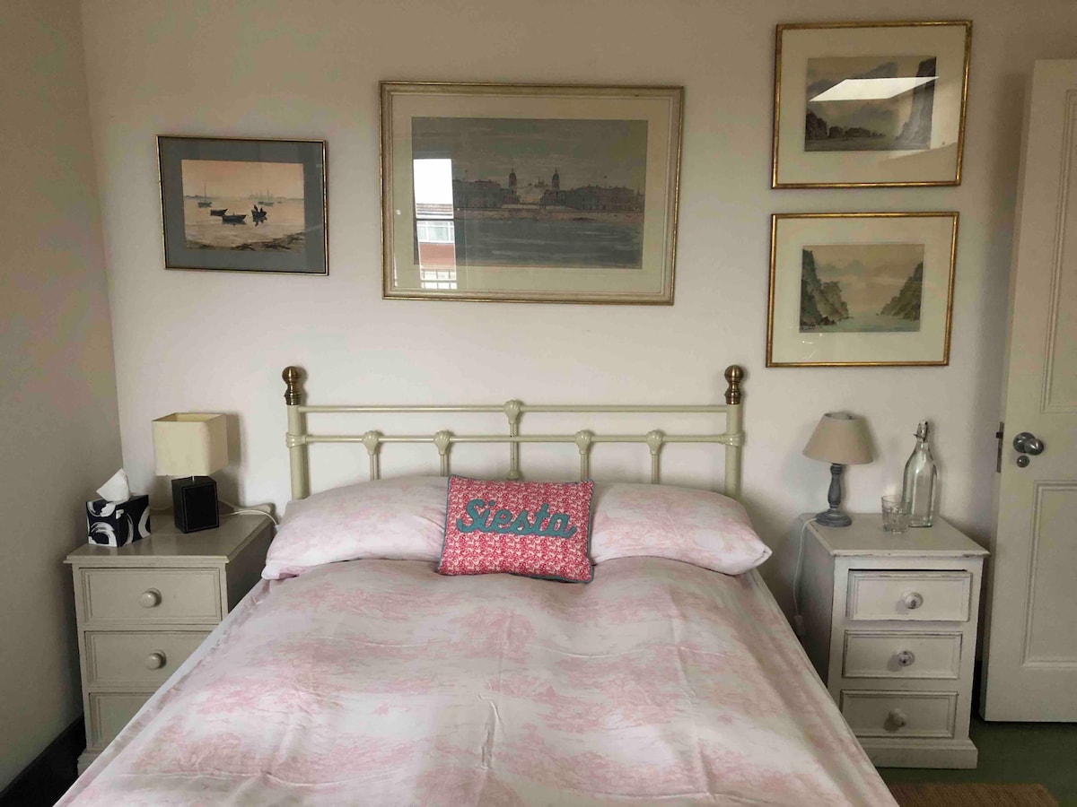A double room is presented with a vintage metal bed frame, adorned with a pink floral coverlet. Each bedside table features a lamp and decorative artwork hangs on the walls, creating a homey environment. Natural light fills the room from a window.