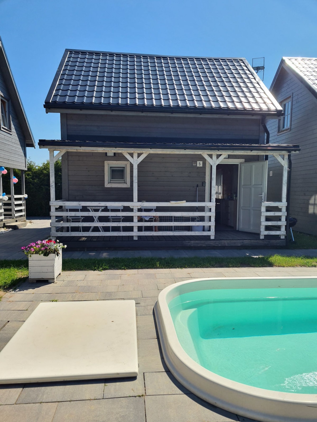 A detached house is presented with a covered porch featuring a seating area. A small pool is visible in the foreground, accompanied by a white storage box. The exterior walls are wooden with a gabled roof, set against a clear blue sky.
