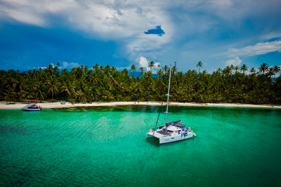 A white catamaran is anchored in clear turquoise waters, surrounded by a lush landscape of palm trees. A pristine sandy beach is visible in the background, with another boat docked nearby under a dramatic sky filled with clouds.