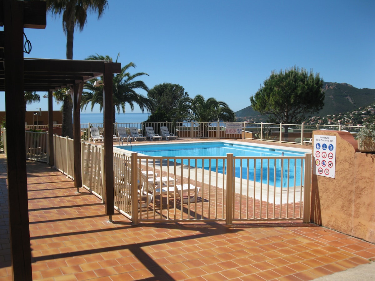 A pool area is enclosed with a white fence, featuring a clear blue swimming pool surrounded by a tiled deck. Several lounge chairs are arranged nearby, and palm trees provide shade. The Mediterranean Sea is visible in the background, adding to the relaxing ambiance.