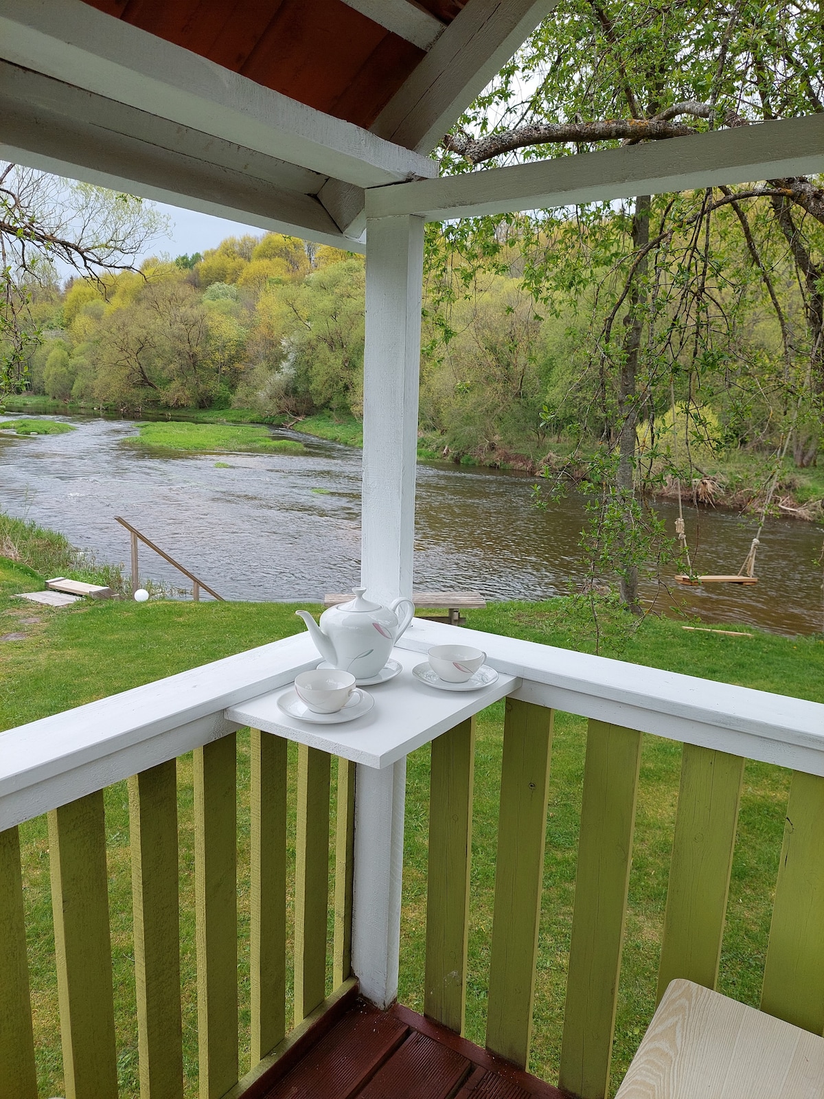 A view from a covered terrace features a white teapot and two cups on a small table. Below, the serene Dubysa river flows, surrounded by lush greenery and trees. A grassy area is visible, leading to a pathway that descends toward the water.