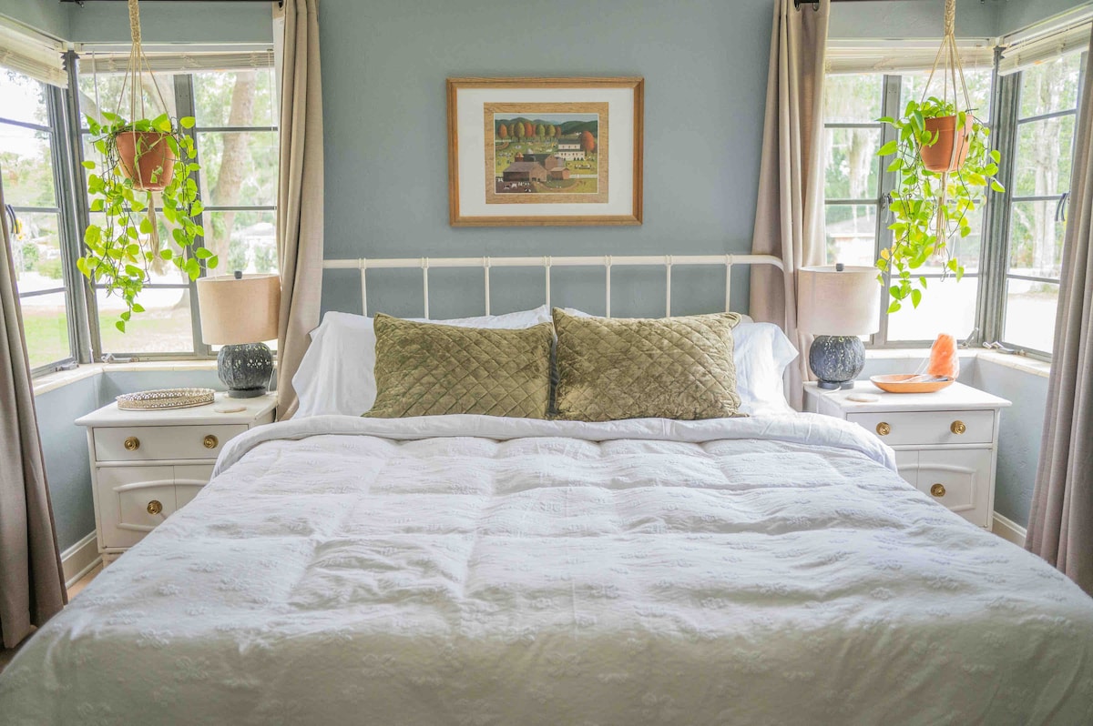The main bedroom features a spacious bed with a white coverlet, accented by two decorative green pillows. Nightstands with lamps are positioned on either side, and plants are suspended from the windows, adding a touch of nature. Light filters through the corner windows.