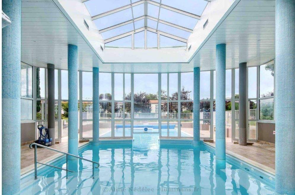 The indoor swimming pool features large glass walls that provide ample natural light. The water appears clear and invites relaxation. Surrounding the pool, tall columns support the high ceiling, creating an open and spacious atmosphere. A view of the outdoor pool area is visible through the glass.