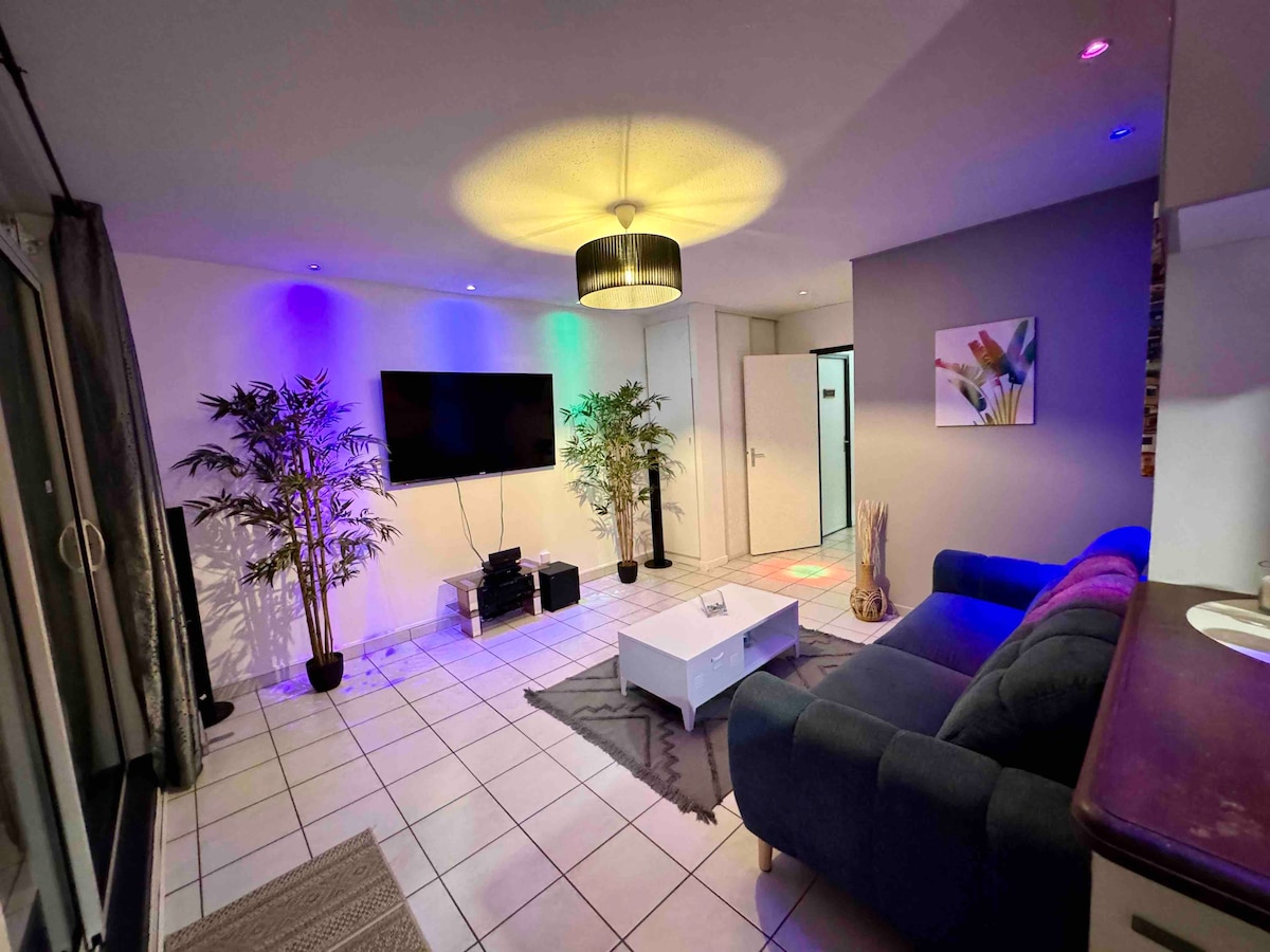 An inviting living space features a dark blue sofa facing a wall-mounted television. Two potted plants add greenery to the room. Soft lighting enhances the atmosphere, while a white coffee table and a textured rug complete the setting. A door leads to additional areas of the property.