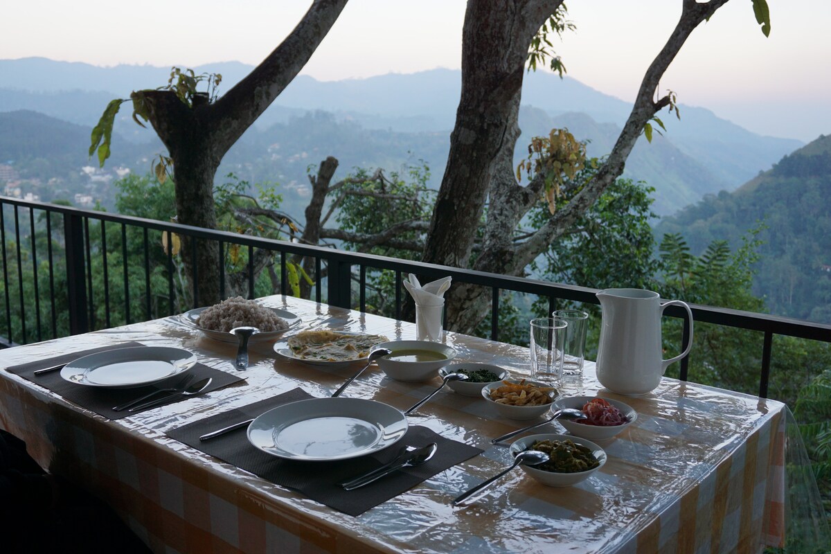 A dining table set on a balcony features white plates and cutlery, alongside a variety of local dishes and condiments. A pitcher and glasses are placed for beverages. The background reveals a scenic view of the mountains and trees, providing a calm and serene atmosphere.
