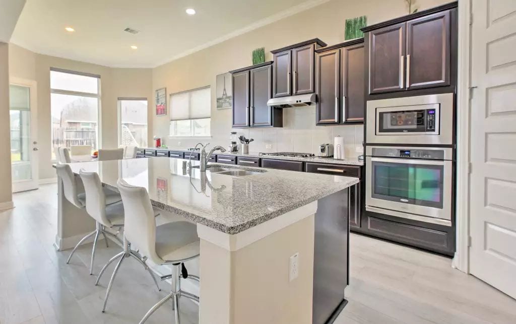 A modern kitchen features dark wood cabinetry and stainless steel appliances. A large granite island provides seating for several guests. Natural light floods in through multiple windows, highlighting the open layout and clean design of the space.