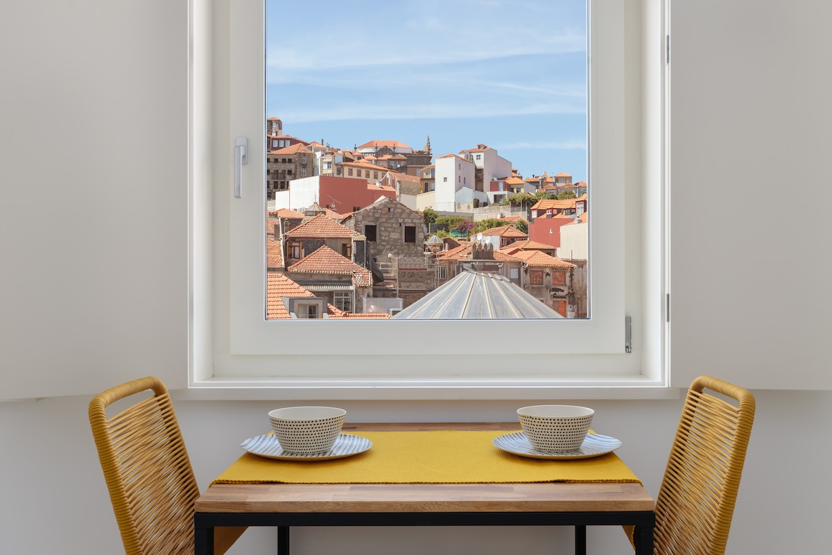A window offers a view of the charming rooftops and architecture of Oporto's Historic Center. A wooden table is set with two bowls on a bright yellow placemat, flanked by two woven chairs, creating a cozy dining area.