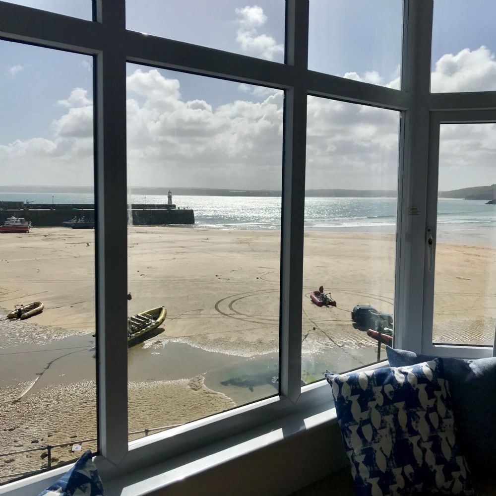 A large window showcases sweeping views of the harbour and bay, capturing boats resting on the sand during low tide. Natural light floods the space, reflecting off the water and highlighting the coastal landscape.