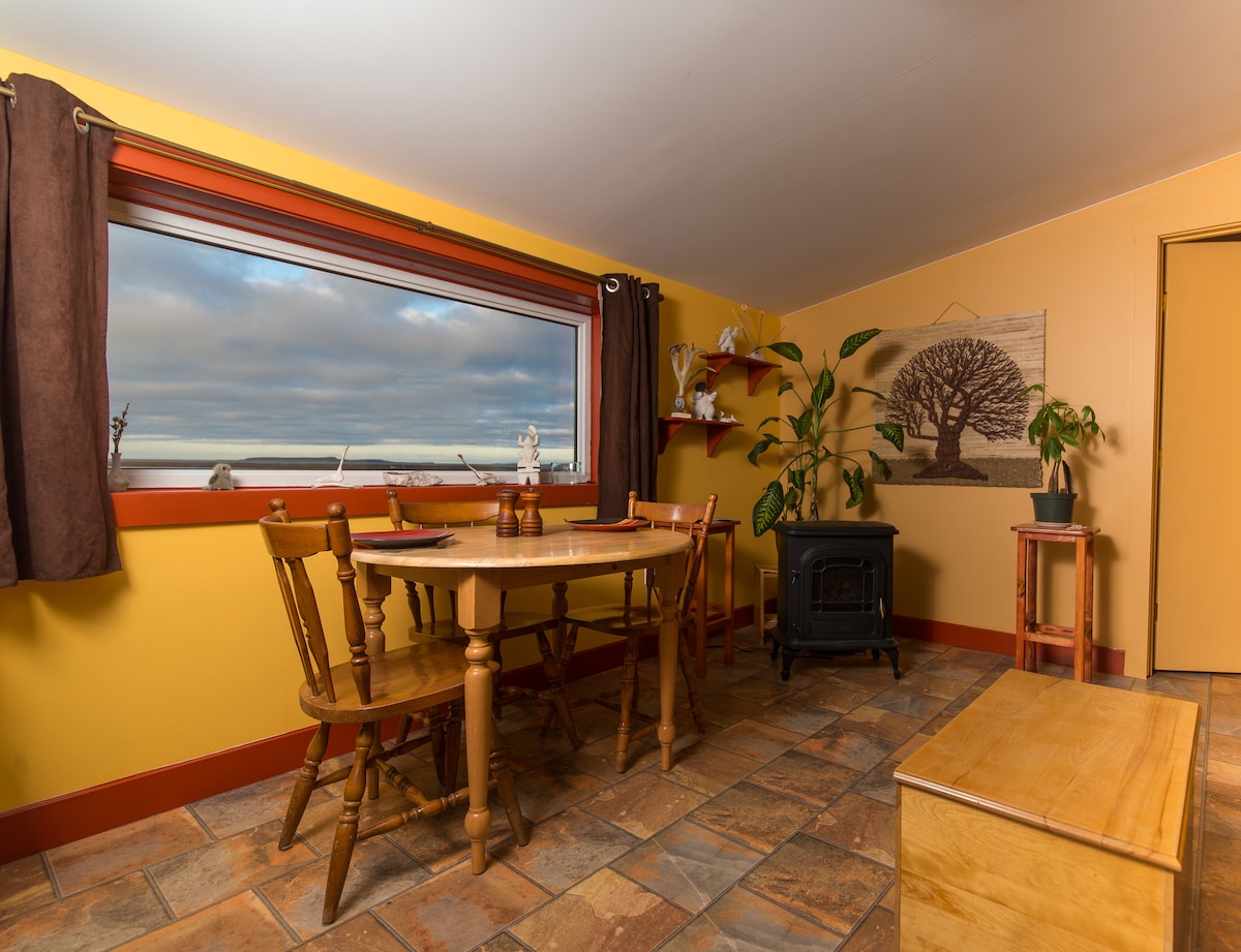 A dining area features a wooden table set with four chairs. A dark wood stove is situated near the table. Large windows provide a view of the landscape, framed by warm-toned walls and dark curtains. Decorative plants add a touch of greenery to the space.