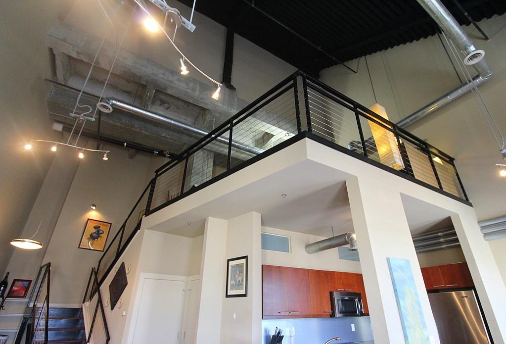 The loft features an open design with a high ceiling, showcasing industrial-style metal railings on the upper level. Warm-toned wood cabinetry is visible in the kitchen area, complemented by stainless steel appliances. Soft lighting from multiple fixtures highlights the spaciousness of the living area.