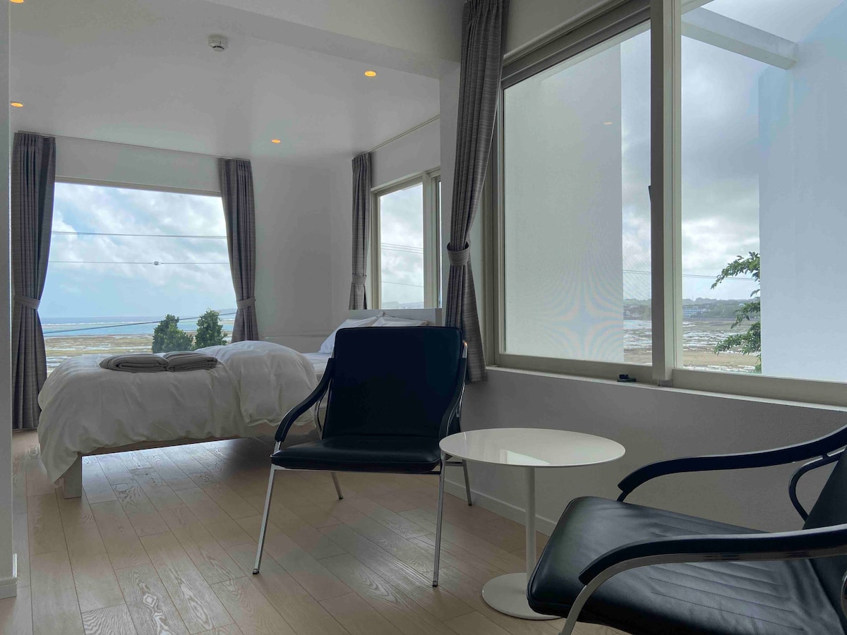 A bright bedroom is furnished with a spacious bed and two contemporary chairs arranged around a small table. Large windows frame the ocean view, allowing natural light to fill the room. Light-colored flooring complements the neutral tones of the decor.