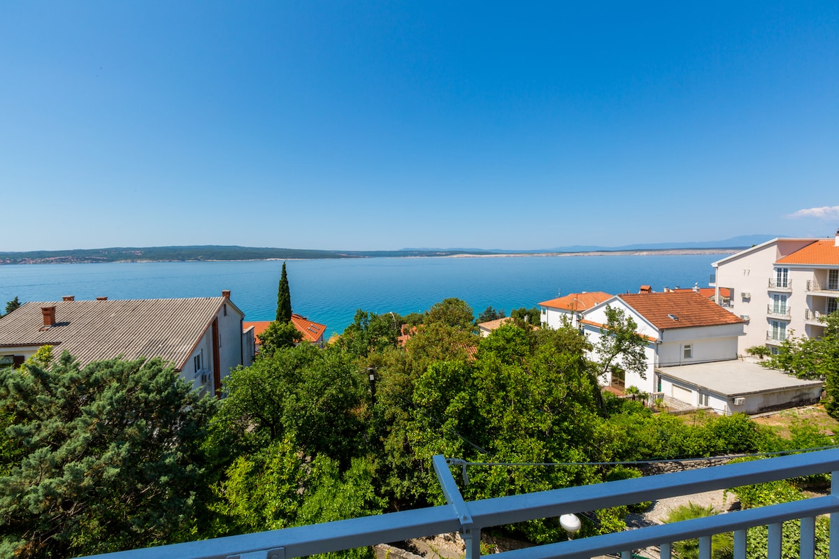A scenic view of the sea is captured from a balcony. The landscape includes lush greenery and residential structures with red roofs, leading to the calm blue water. The clear sky enhances the serene atmosphere of this coastal location.