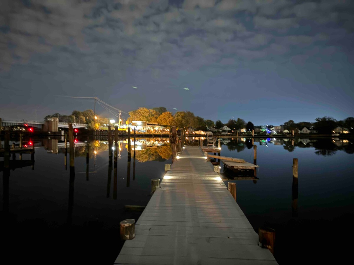A private dock extends over calm waters, illuminated by subtle lighting along the walkway. Reflections of trees and distant lights create a serene atmosphere under a night sky filled with soft clouds.