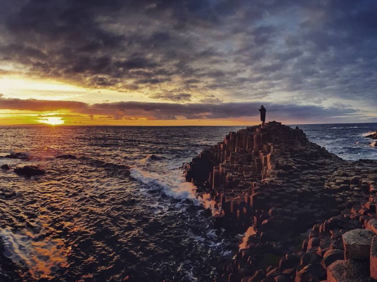 A rocky coastal formation rises from the sea, surrounded by waves. A person stands on top, silhouetted against a vibrant sunset that casts warm hues over the horizon. Dramatic clouds fill the sky, enhancing the natural beauty of the scene.