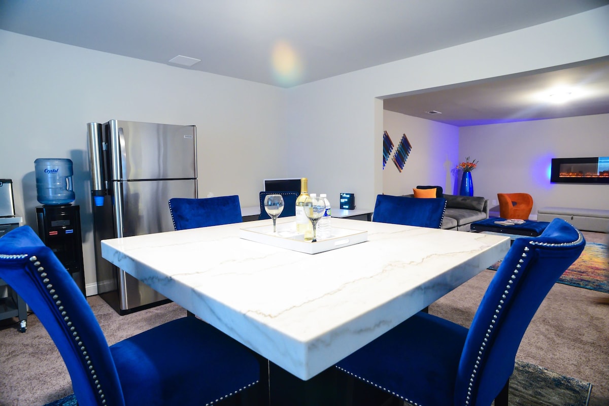 A modern dining area features a marble-top table surrounded by plush blue chairs. A stainless steel refrigerator and a water cooler are positioned nearby. The open layout leads to a cozy living area, where a soft seating arrangement is visible in the background.