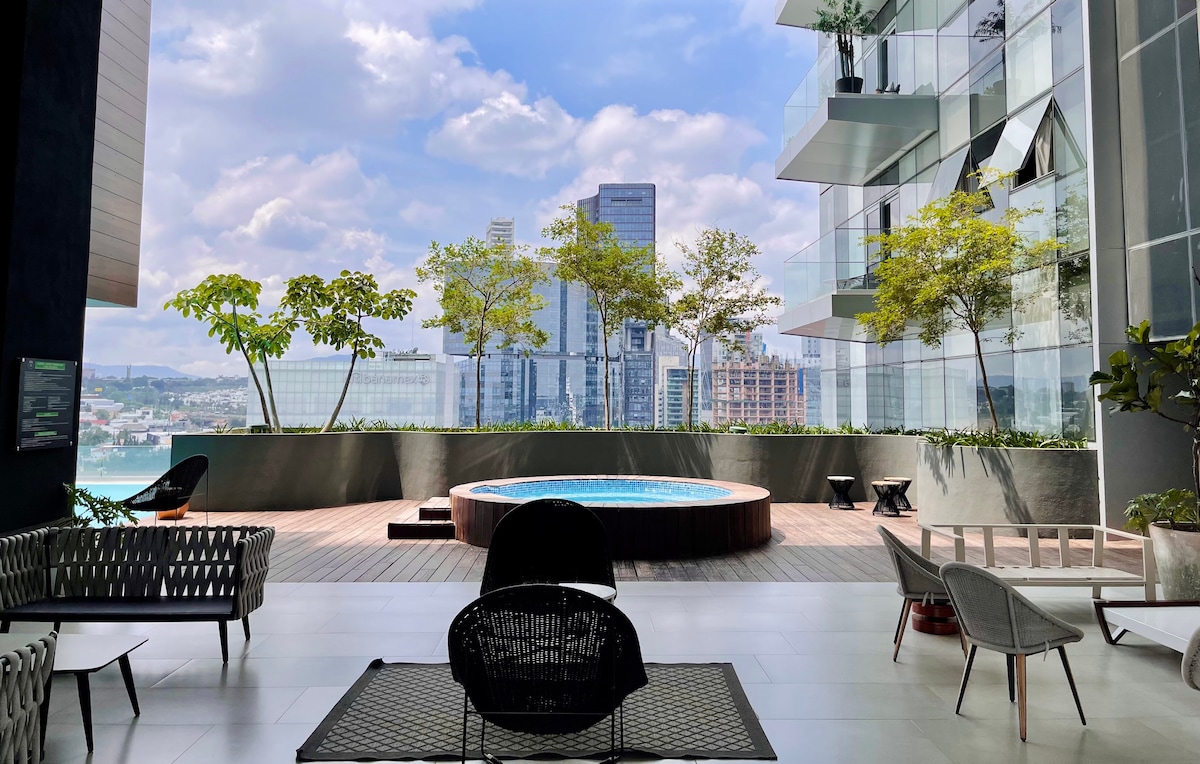 A spacious outdoor lounge area features modern seating arrangements with a circular hot tub in the background. Lush greenery and city skyline views create a relaxing atmosphere, while large glass windows enhance the brightness of the space.