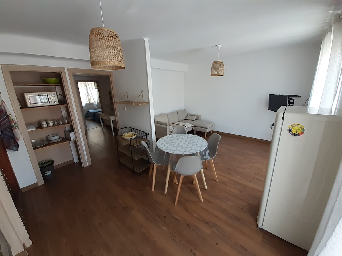 A bright living area features light wooden flooring and a relaxed atmosphere. A round dining table with chairs is positioned near the open kitchen, while a comfortable sofa occupies one side. A flat-screen television is mounted on the wall, and natural light enters through large windows.