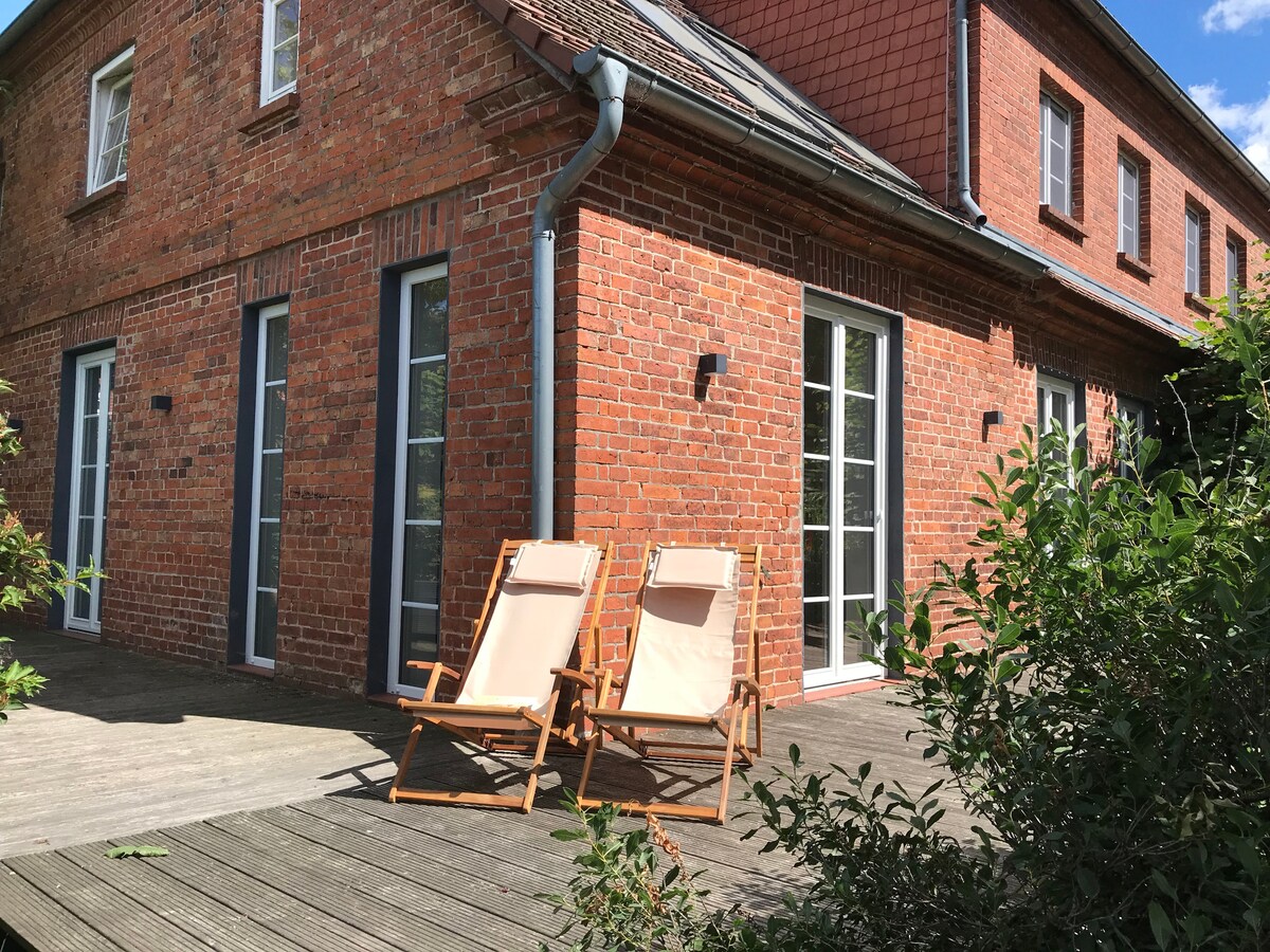 A sunlit terrace features two wooden chairs, inviting relaxation. The red brick facade of the house adds character, with multiple glass doors providing an airy connection to the outdoors. Surrounding greenery enhances the serene outdoor setting, creating spaces for open-air enjoyment.