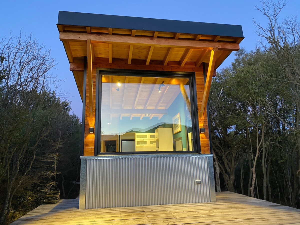 The tiny home features a modern wooden exterior with large glass windows, offering a clear view of the interior. The structure is elevated on a wooden deck, surrounded by native trees, highlighting a connection to nature while being well-lit from above.