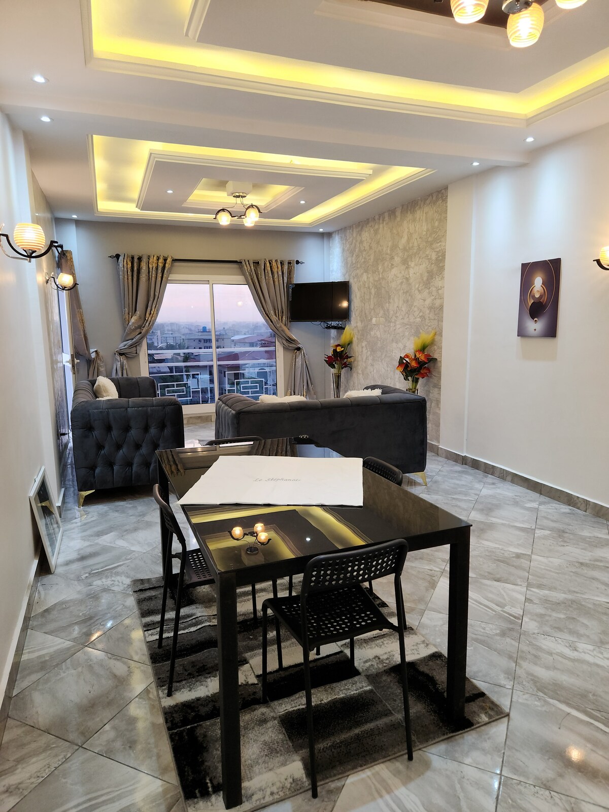 A modern living space is highlighted by a sleek table with black chairs, surrounded by plush seating. Soft lighting from the ceiling enhances the ambiance, while large windows offer a view of the skyline. Decorative plants add a touch of color to the room.