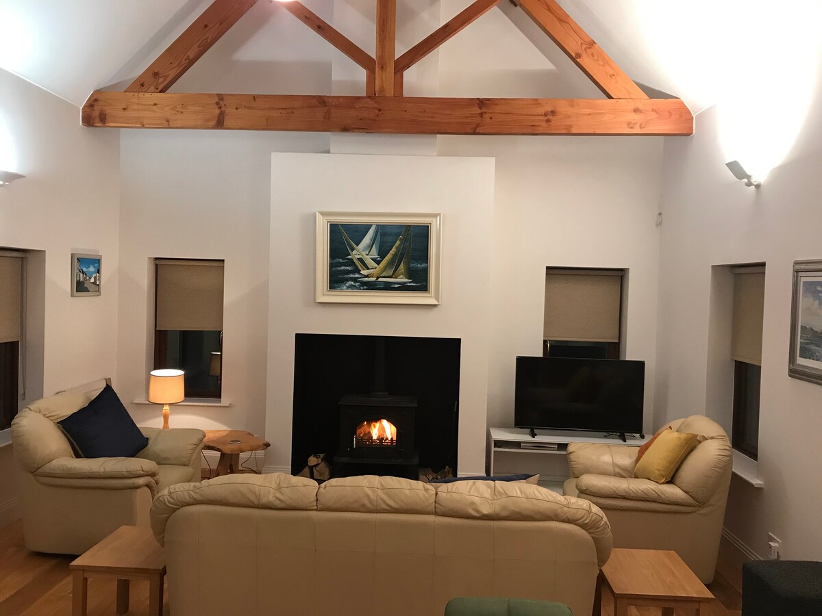 A spacious living room features a high ceiling supported by exposed wooden beams. Two cream-colored sofas are arranged around a central coffee table, facing a fireplace. A television stands beside the fireplace, and soft lighting enhances the cozy setting.
