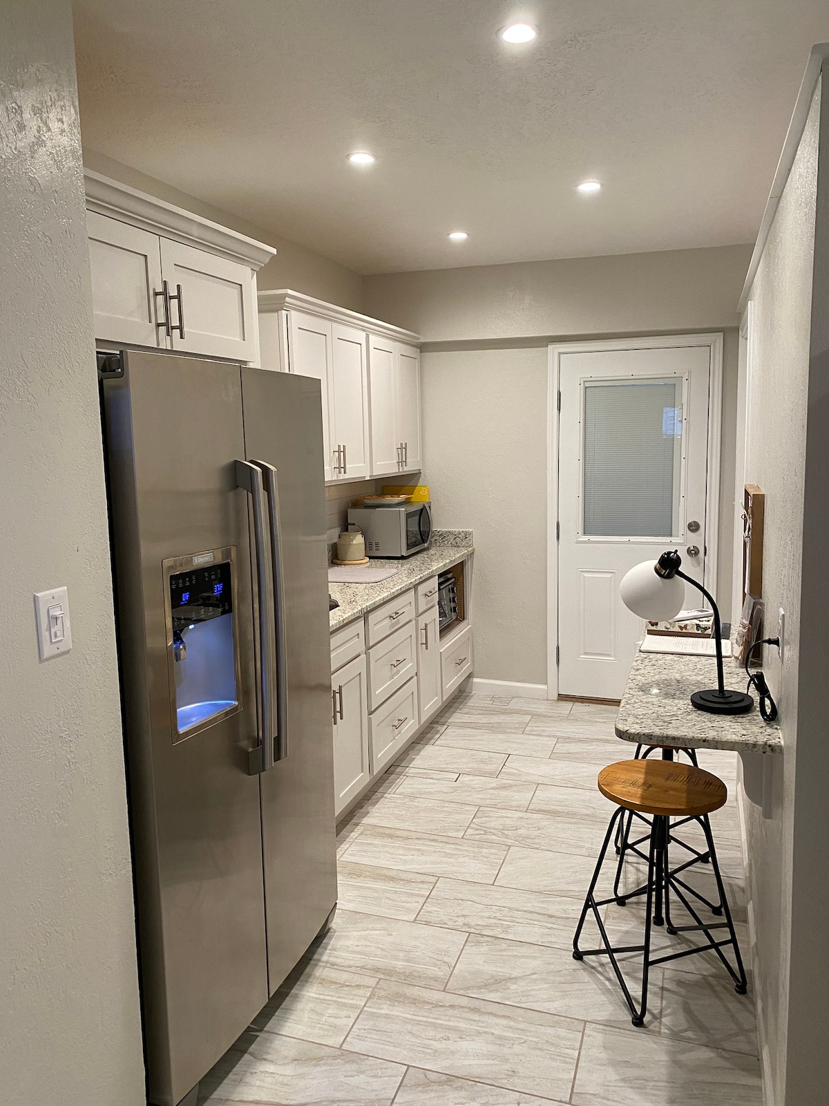 A modern kitchenette is featured, equipped with stainless steel appliances, including a large refrigerator. Bright white cabinetry complements the light-colored countertops. A small workspace with a bar stool is positioned by the window, which provides natural light and a view of the outdoors.