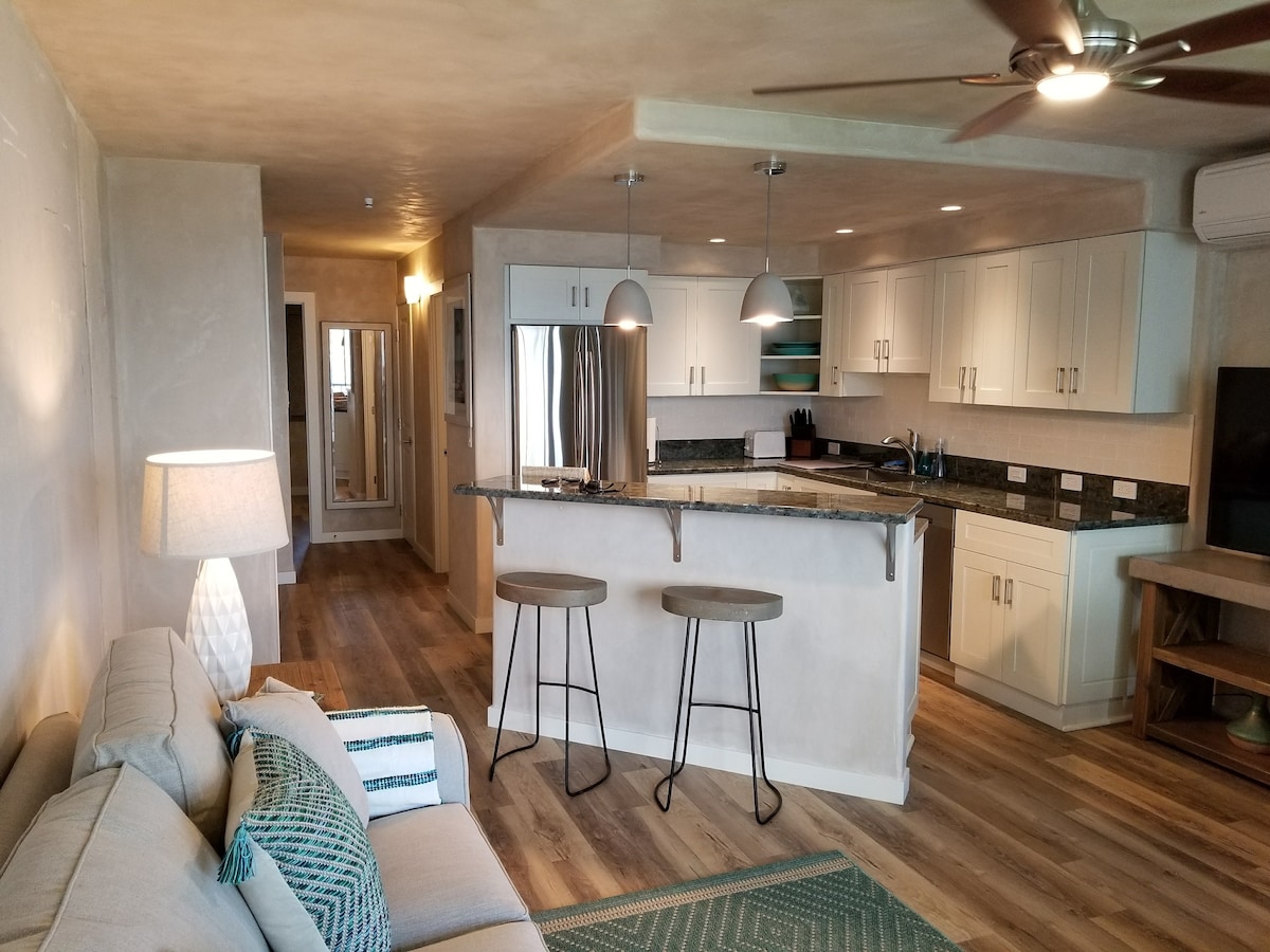 An open living area showcases a comfortable sofa and a textured area rug. A modern kitchen features stainless steel appliances and a granite countertop, complemented by two barstools. Soft lighting from pendant lamps and a nearby floor lamp enhances the inviting ambiance.