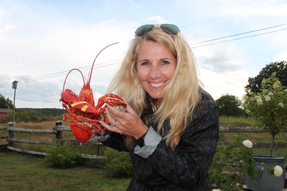 A person holds a large lobster in an outdoor setting, with fields and trees visible in the background. The scene is framed by a rustic wooden fence and greenery, suggesting a relaxed atmosphere on the farm.