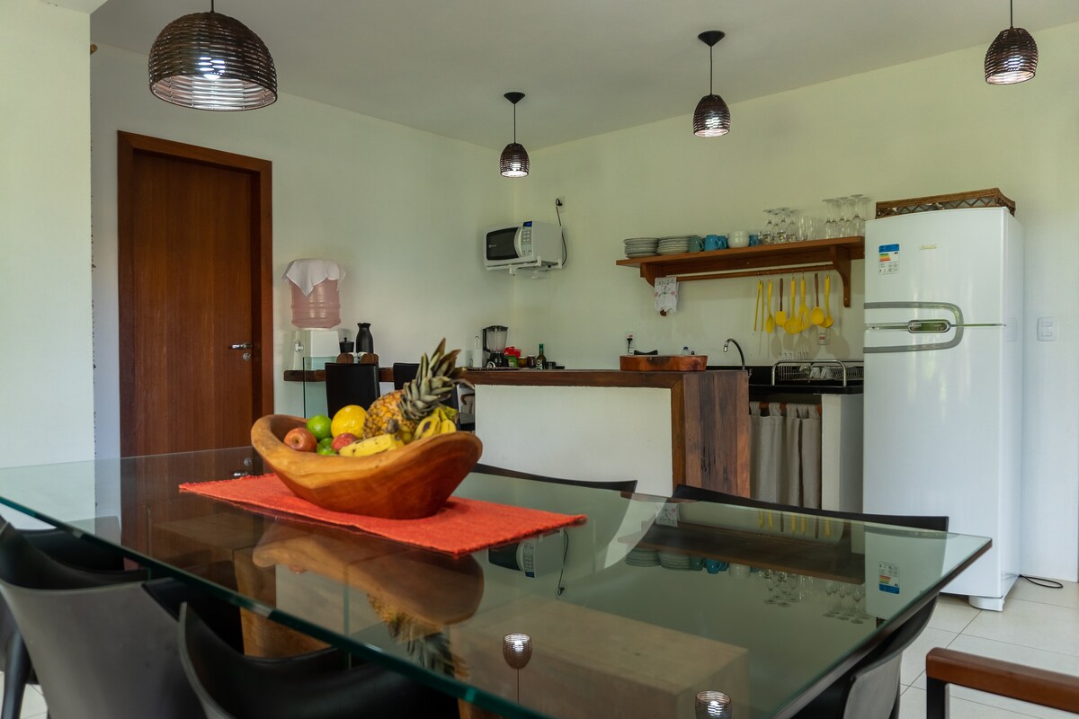 A dining area features a glass table surrounded by black chairs, with a wooden bowl of fruit at the center. An open kitchen is visible in the background, equipped with appliances including a refrigerator and a coffee maker, illuminated by pendant lights.