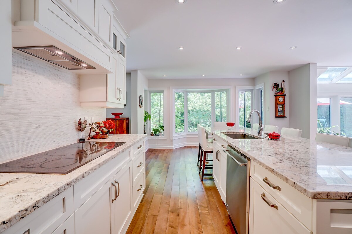 A modern kitchen showcases elegant white cabinetry and a spacious marble countertop. High-end appliances are positioned for convenience. Natural light floods in through large windows, illuminating the open layout, which features hardwood flooring and inviting spaces for family gatherings.