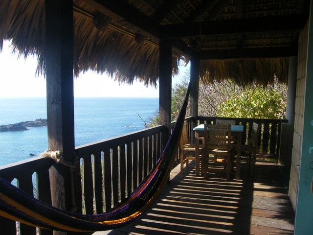 A covered terrace offers stunning ocean views, featuring a wooden dining table with chairs and a colorful hammock. Sunlight creates a pattern of shadows on the wooden floor, enhancing the rustic charm of this peaceful outdoor space.