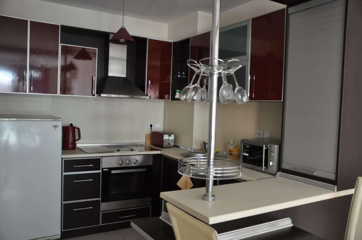 A modern kitchen is showcased with sleek cabinetry in dark and light tones. Built-in appliances, including an oven, microwave, and refrigerator, are visible. A countertop with seating is present, complemented by a hanging rack for glasses. Natural light filters through a window covered by blinds.