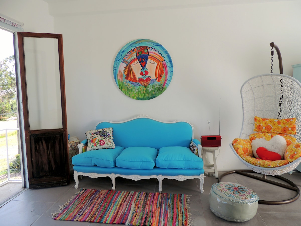 A bright living area is depicted, featuring a vibrant blue sofa complemented by a multi-colored area rug. A decorative wall hanging adds artistic flair, while a unique hanging chair with colorful cushions invites relaxation. Natural light streams in through an open doorway.