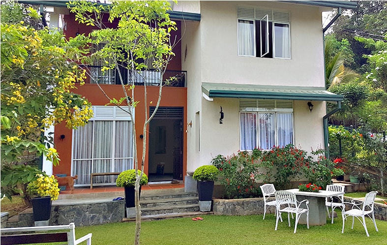 A two-story bungalow is set in a well-maintained garden, featuring colorful flowers and manicured greenery. The exterior includes sliding glass doors and large windows, ensuring ample natural light. Outdoor seating is provided with white chairs and small tables, inviting relaxation in the tranquil surroundings.