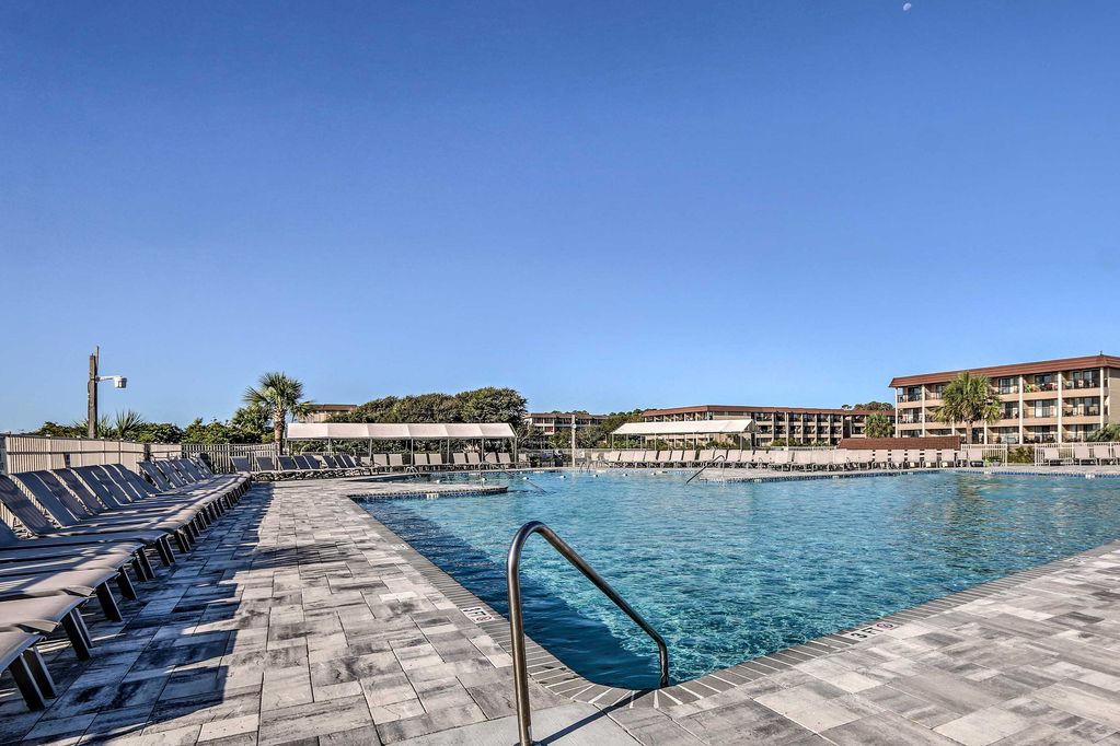 The spacious outdoor pool area is surrounded by lounge chairs arranged neatly along the perimeter. Lush palm trees provide subtle greenery in the background, while buildings of the resort stand at a distance against a clear blue sky.
