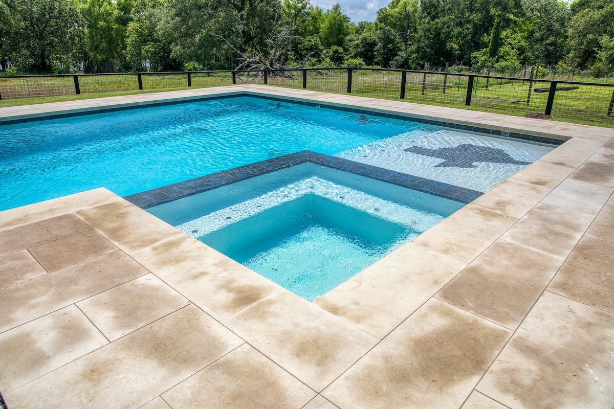 A large lap pool is framed by a smooth stone deck, showcasing clear blue water. A spa section is integrated into the pool design, creating an inviting space for relaxation. Surrounding greenery is visible beyond the pool area, offering a sense of nature and tranquility.