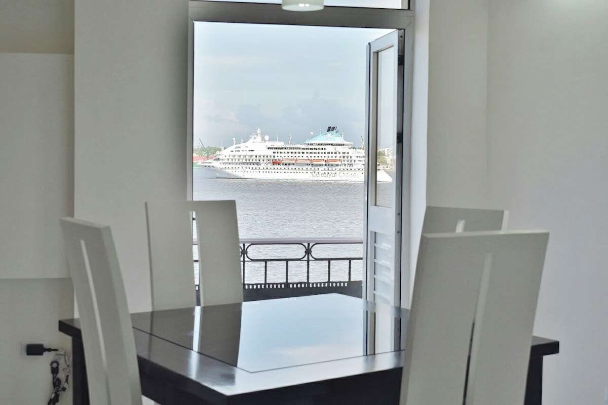 A view from the apartment showcases a large ship in the bay, visible through an open window. The image features a black dining table with four white chairs, positioned near the window. Natural light illuminates the space, enhancing the airy atmosphere.