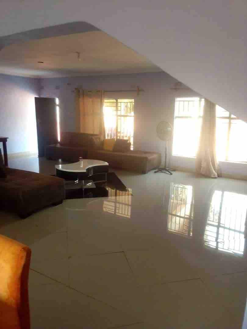 A spacious living room features a combination of brown sofas arranged around a light coffee table, with a smooth tiled floor reflecting natural light. Large windows are adorned with light curtains, allowing soft illumination to enhance the room's openness.