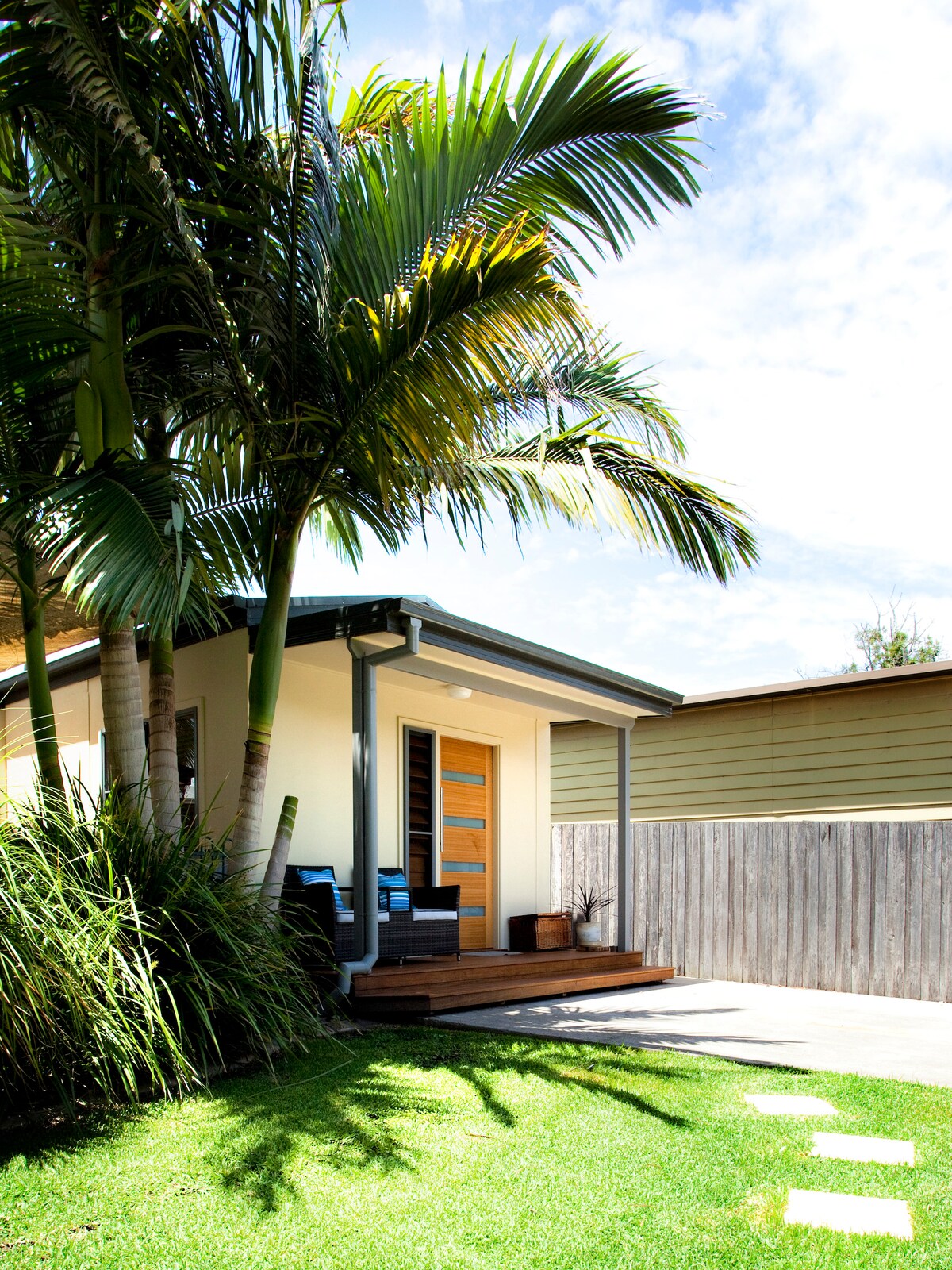 The exterior of a stylish villa features a welcoming entryway with modern wooden doors. Comfortable seating is positioned on a small covered porch, surrounded by vibrant greenery and palm trees. A pathway leads to the entrance, with a fence providing privacy.