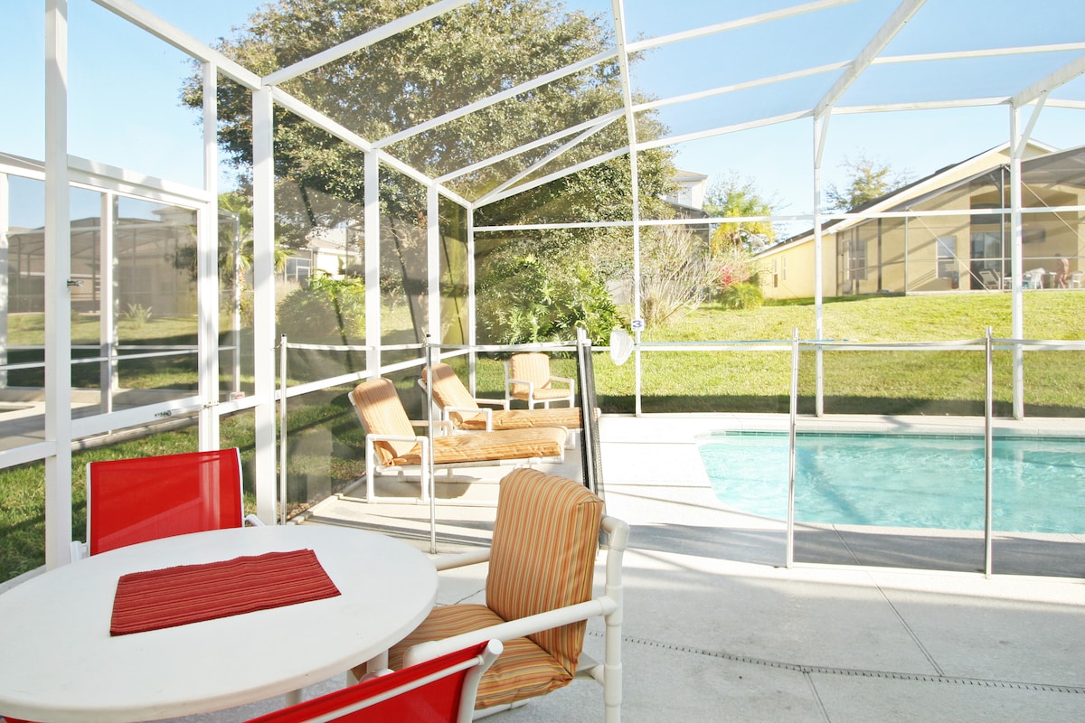 A screened lanai surrounds a private in-ground pool, offering seating options including lounge chairs and a dining table. Sunlight filters through the enclosure, highlighting the tranquil outdoor space and lush greenery in the background.