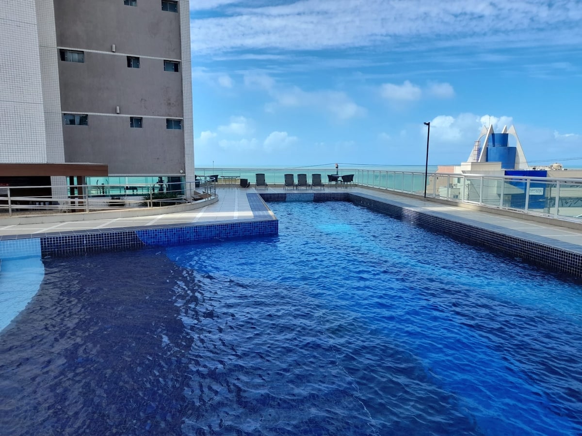 An outdoor pool reflects the clear blue sky, surrounded by a spacious deck with lounge chairs. The pool's surface shimmers in various shades of blue, with a glass railing providing a view of the surrounding area, including buildings in the distance.