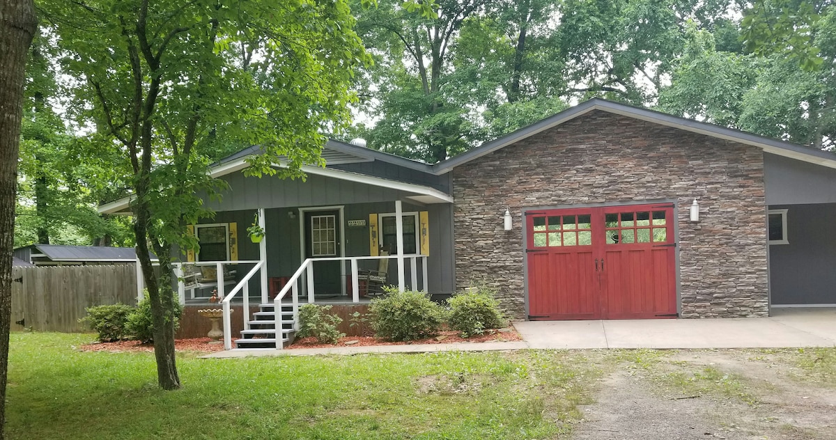 A charming house is framed by lush green trees, featuring a combination of horizontal siding and stone accents. A welcoming entrance is highlighted by a set of stairs leading to the front porch, and a two-car garage with red doors is visible to the right.
