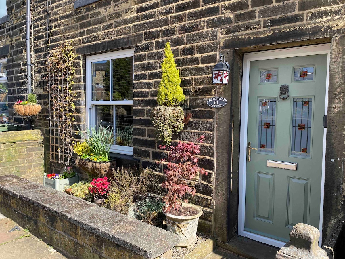 The entrance of Maple Leaf Cottage is framed by a charming stone facade and features a light green door with decorative stained glass. Flower pots filled with vibrant plants and a small stone wall enhance the welcoming atmosphere.