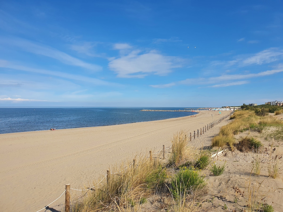 A broad sandy beach extends alongside the gently lapping waves of the ocean, framed by sparse grass and wooden fencing. In the distance, sunbathers can be seen on the sand, while beach umbrellas are positioned further along the shore.