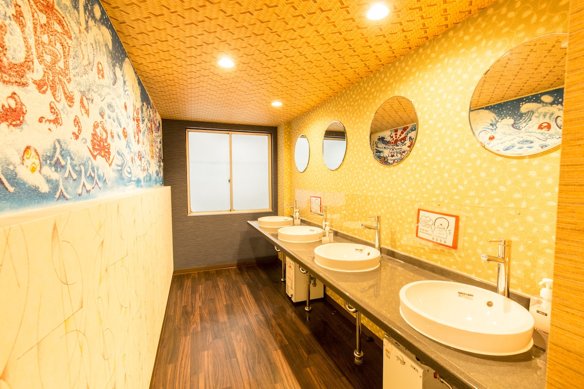 The bathroom features multiple sinks set against a detailed mural backdrop. Bright circular mirrors above the sinks complement the warm-toned walls. A large window allows natural light to enter, enhancing the spacious atmosphere of the area.