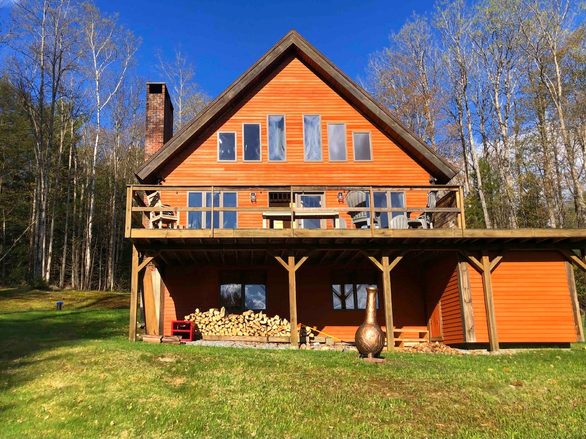 The chalet displays a warm orange exterior with multiple large windows. A spacious deck extends across the front, featuring outdoor seating options. A woodpile is neatly stacked below the deck, alongside a decorative outdoor feature. Surrounding trees provide a natural backdrop.