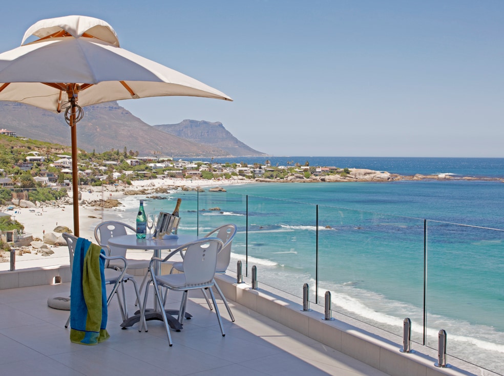 A balcony is showcased, featuring a round table set with chairs, and an umbrella providing shade. Clear glass railings offer unobstructed views of the turquoise ocean and sandy beach below. Lush mountains and a blue sky create a serene backdrop for relaxation.