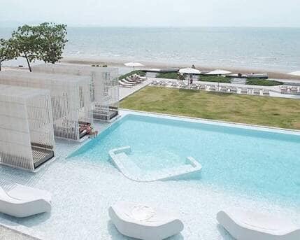 A clear view of a beachfront pool features gentle waves lapping at the shore beyond. Several white sun loungers are positioned around the pool, with shaded seating areas visible nearby. A well-maintained lawn complements the serene environment, inviting relaxation by the ocean.